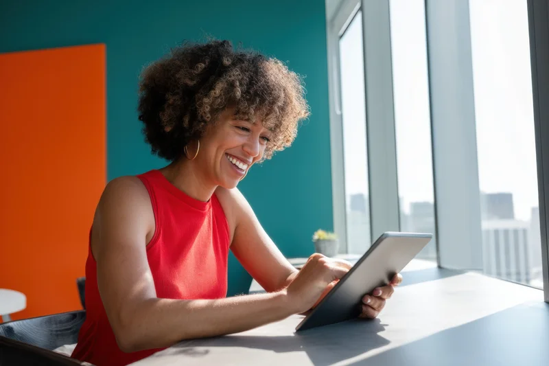 Woman smiling while using tablet at desk by window in modern office