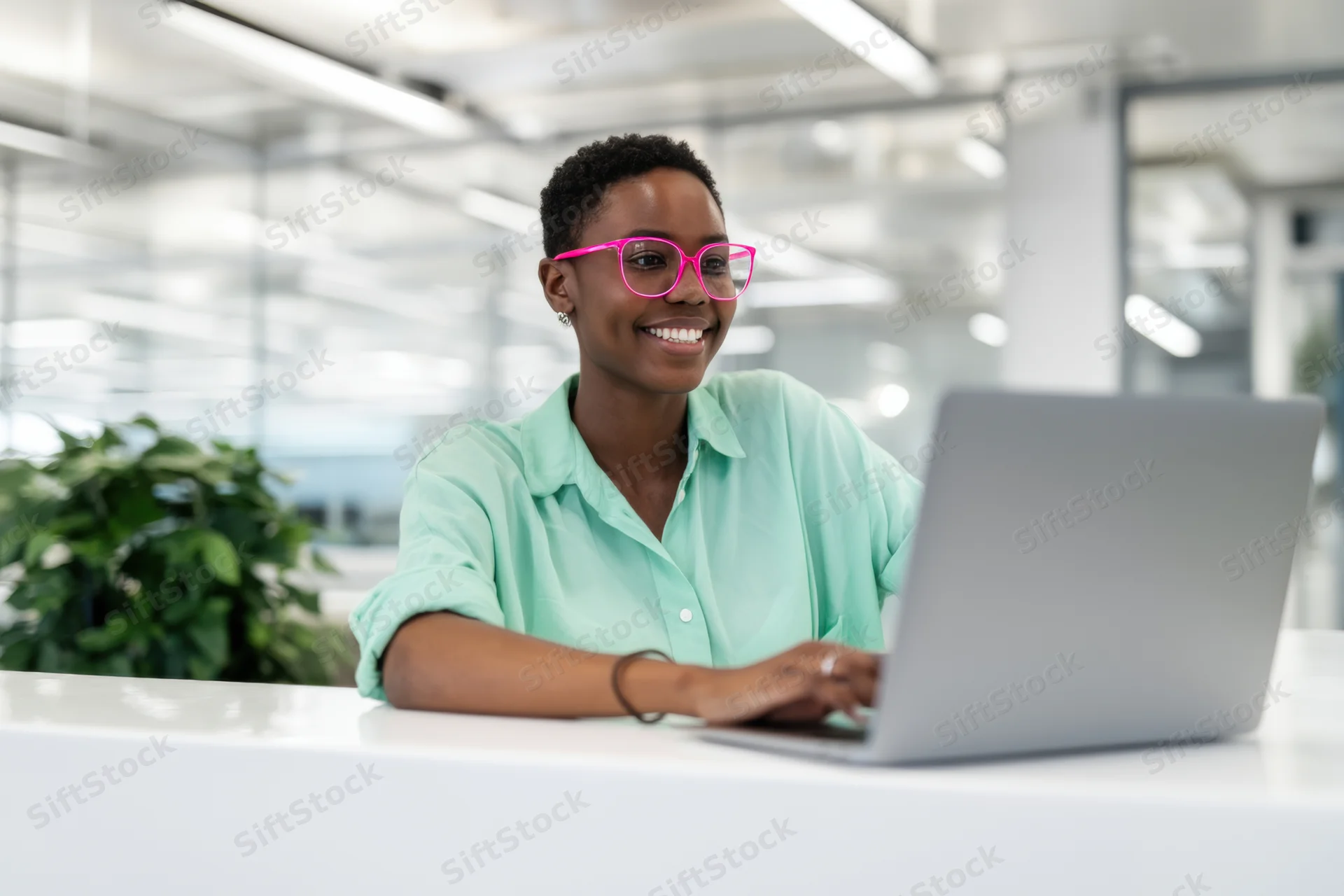 Smiling woman in pink glasses working on laptop at office desk