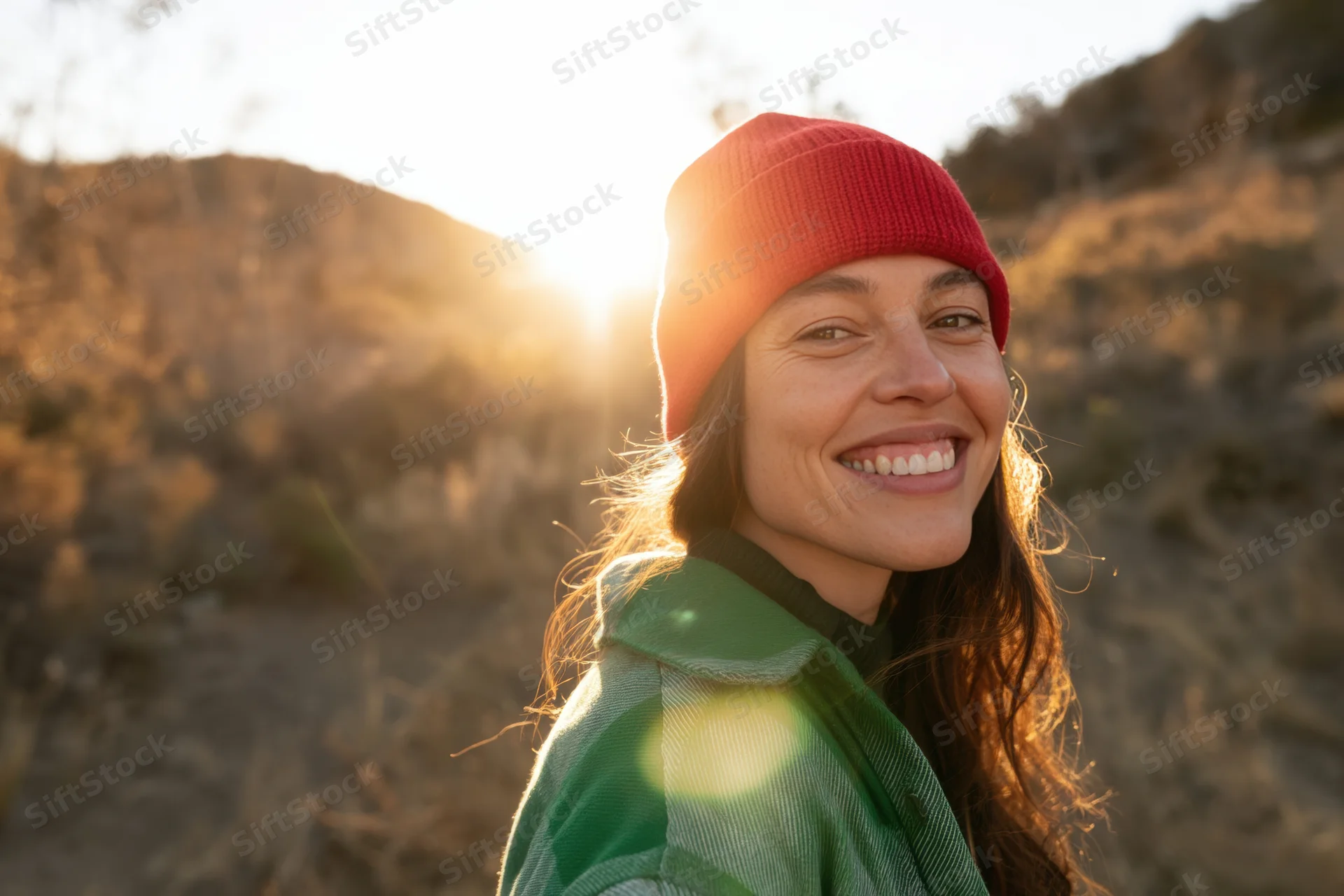 Smiling young woman outdoors in golden hour light wearing a red beanie and green jacket, surrounded by dry hills and warm sunlight