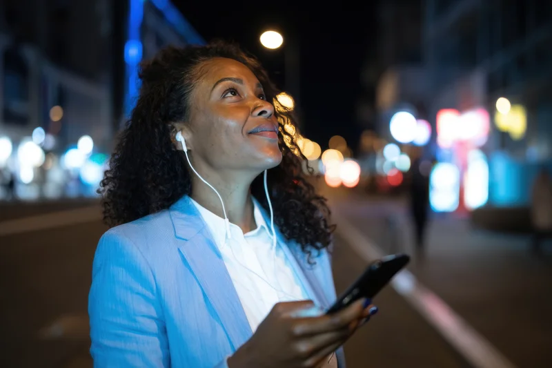 Woman in light blue blazer holding smartphone with earphones, smiling while walking on city street at night