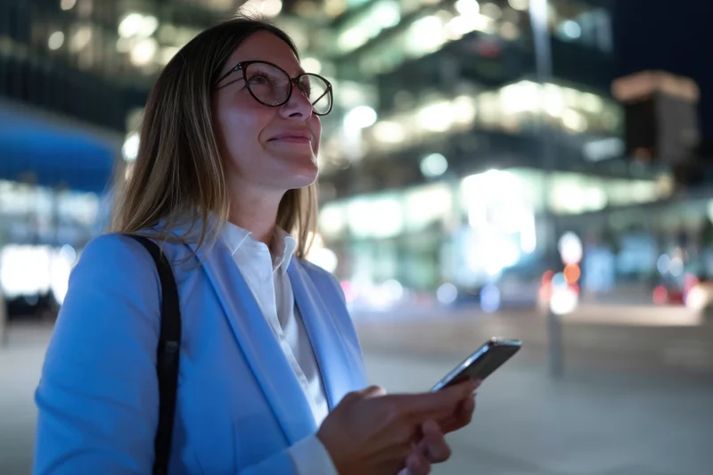 Woman in light blue blazer holding smartphone, smiling, standing in city at night