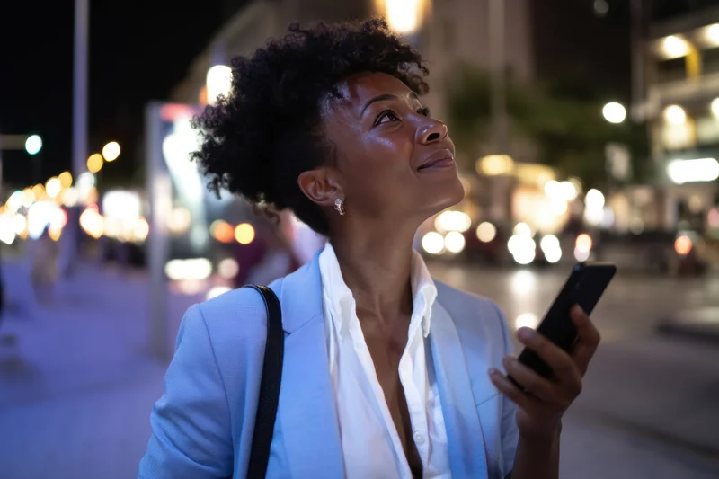 Woman in light blue blazer holding smartphone while looking up on city street at night