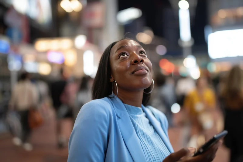 Woman in light blue blazer holding smartphone and looking up in a bustling city street at night