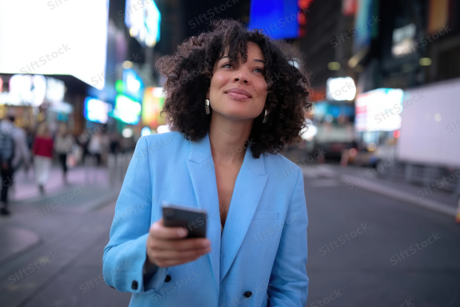 Woman in light blue blazer holding smartphone on bustling city street at night