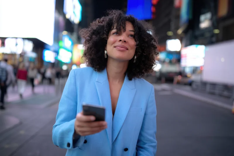 Woman in light blue blazer holding smartphone on bustling city street at night