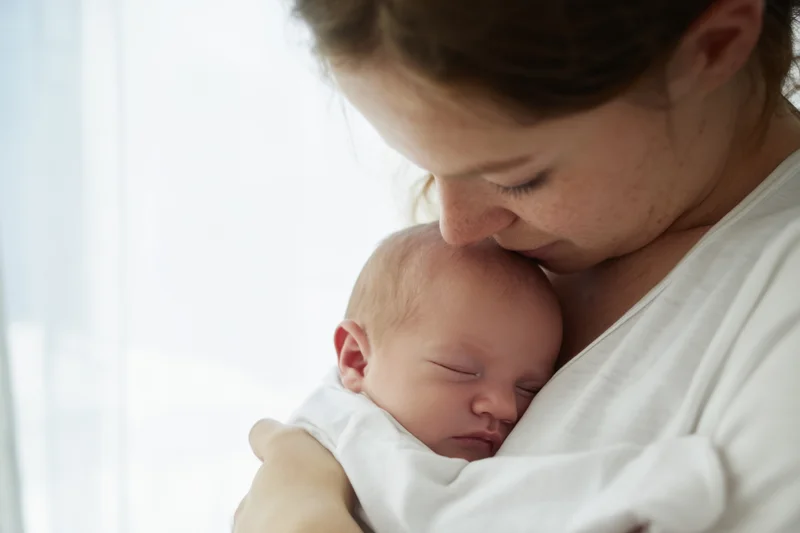 Mother cradling sleeping newborn baby close to her chest indoors near window