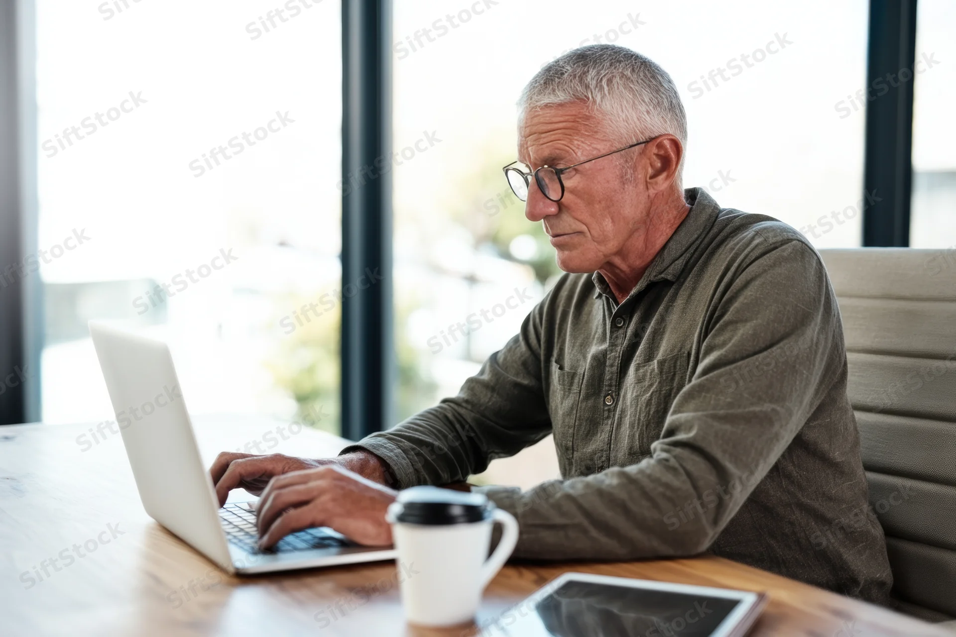Senior man typing on laptop at wooden desk in bright office space