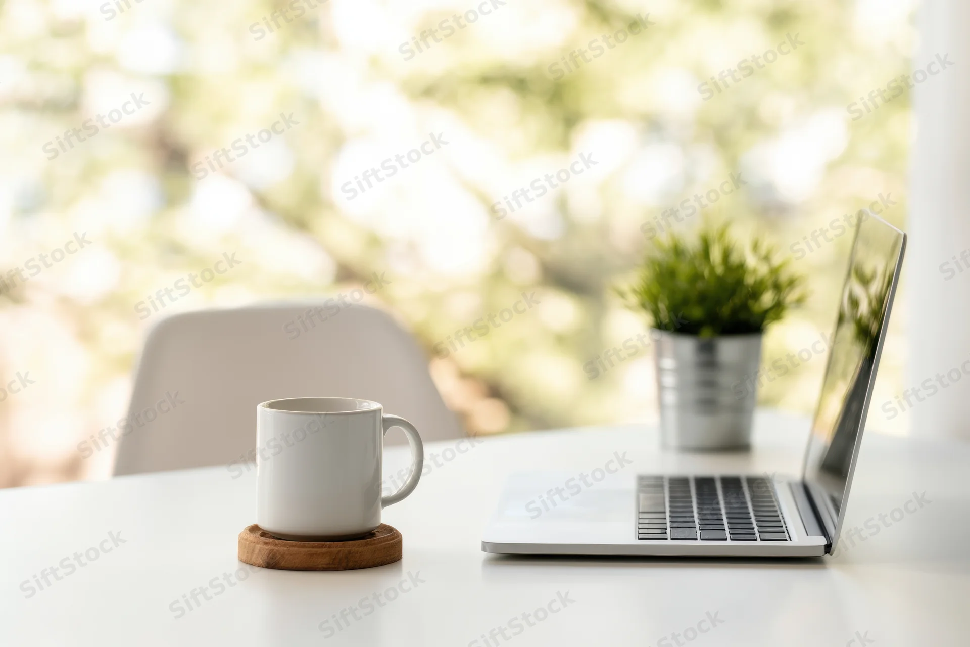 Silver laptop open on white desk beside white mug on wooden coaster with small potted plant, natural light from window in background