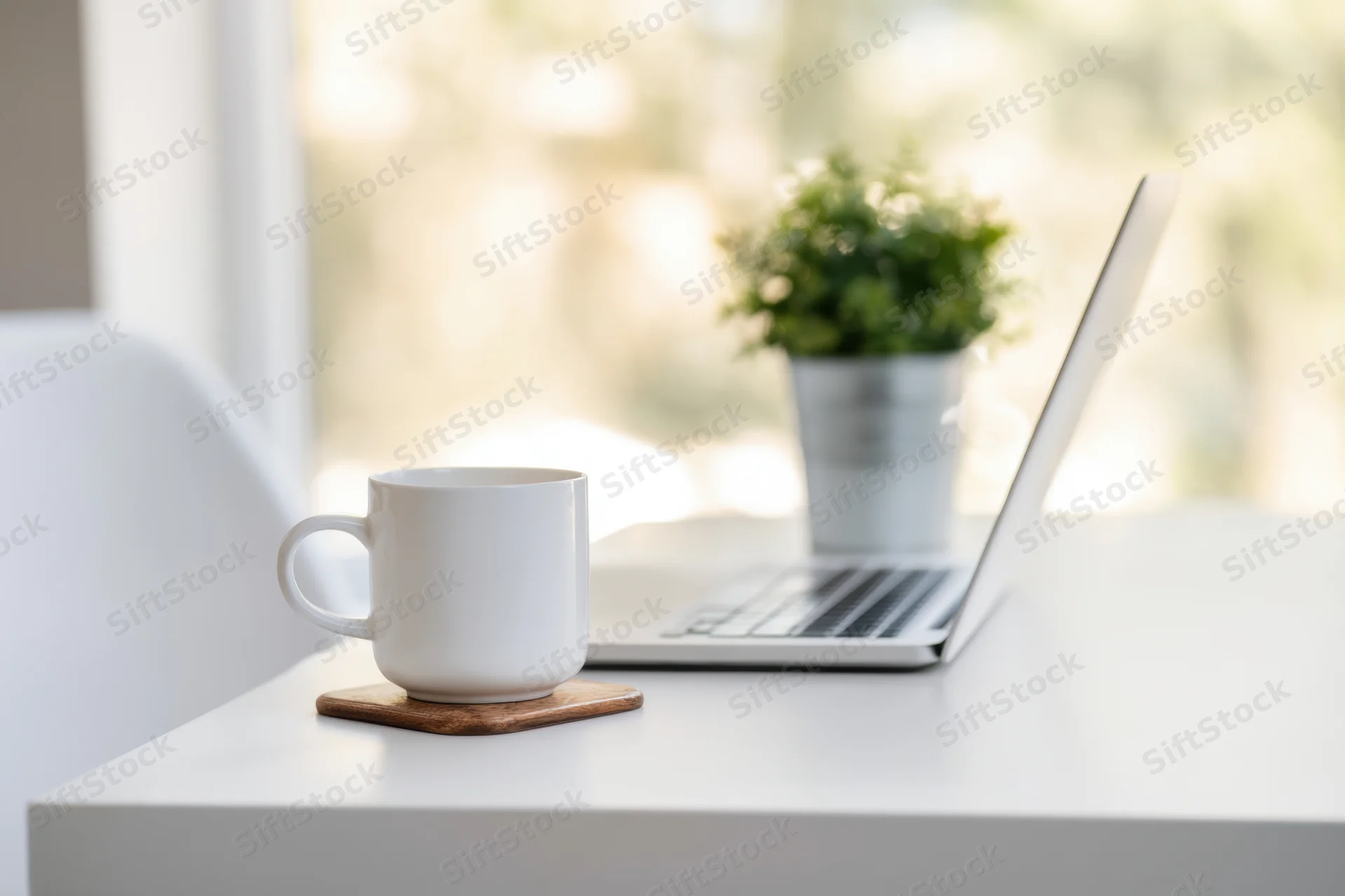 Slim silver laptop open on white desk beside white mug on wooden coaster with potted plant near window