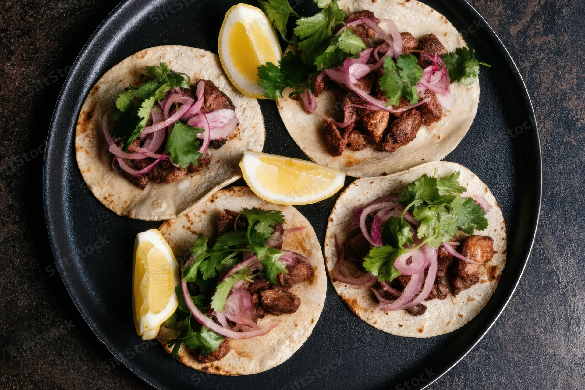 Four tacos with meat, cilantro, red onion and lemon wedges arranged on a black plate