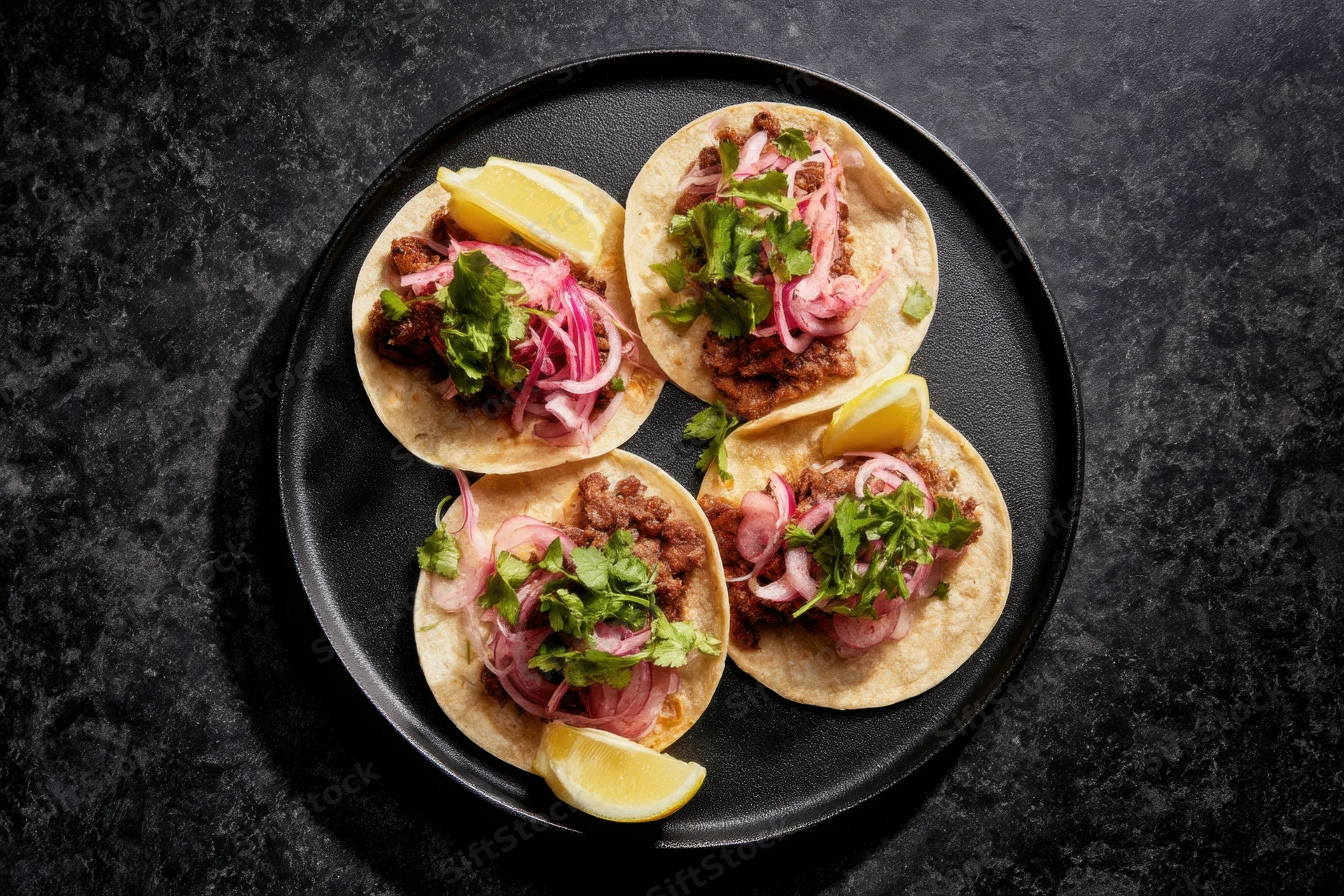 Four tacos with meat filling, cilantro, pickled onions and lemon wedges arranged on a round black plate viewed from above