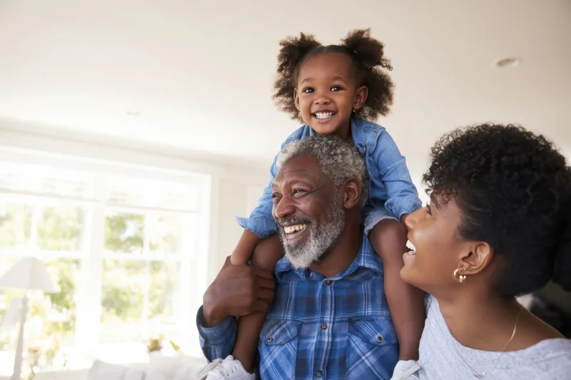 Black multi-generational family smiling together in sunlit living room with child on father's shoulders