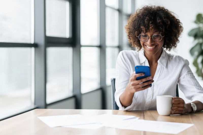 Woman smiling while using smartphone at office desk by large window