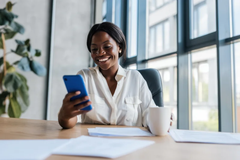 Woman smiling while using smartphone at wooden desk in modern office by large window