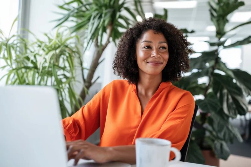 Woman in orange blouse smiling while typing on laptop at desk in office with plants