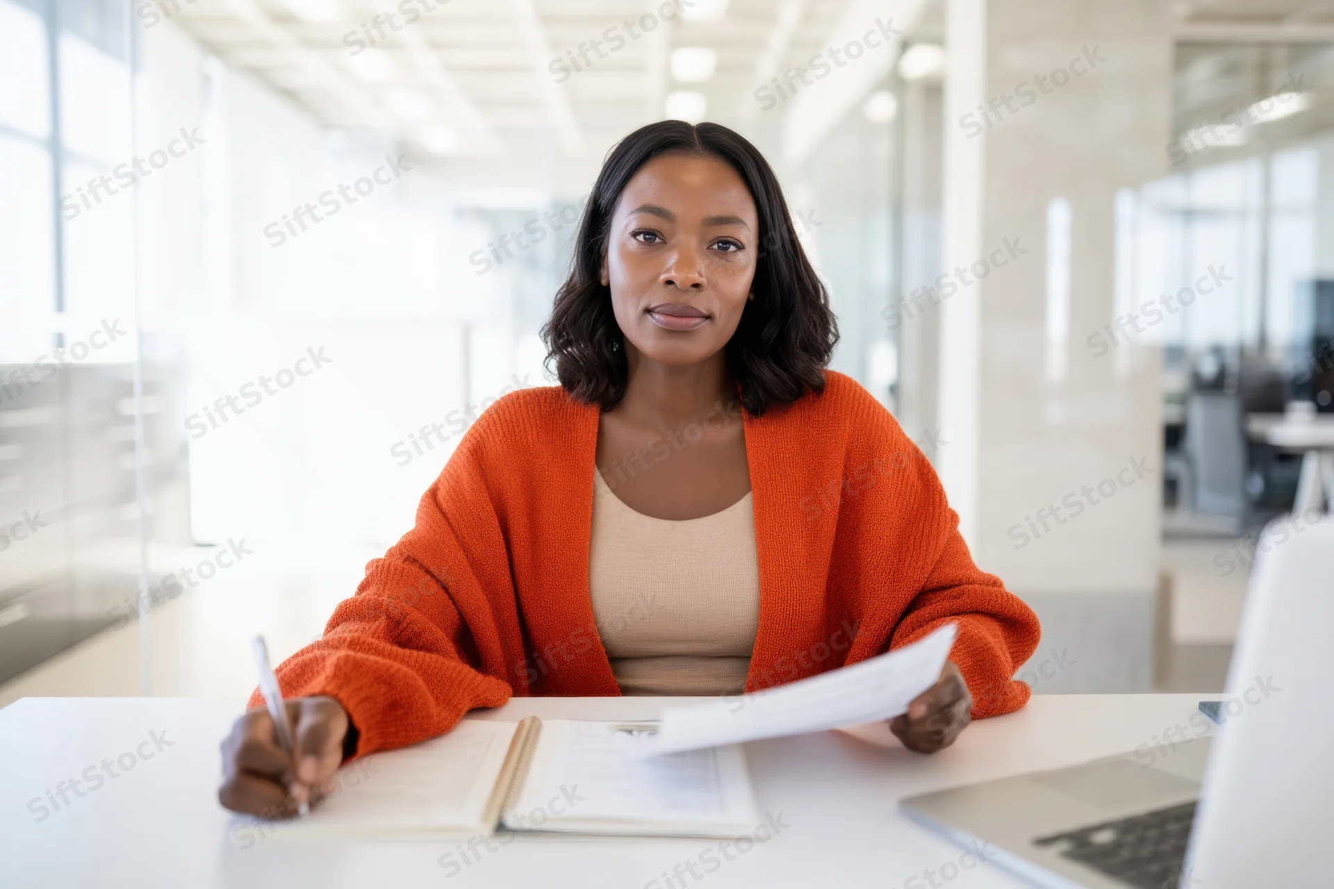 Woman sitting at office desk holding paper and pen with laptop nearby