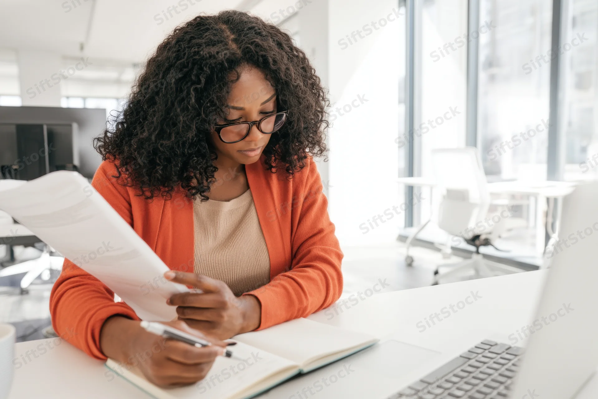 Woman reviewing document while writing in notebook at modern office desk