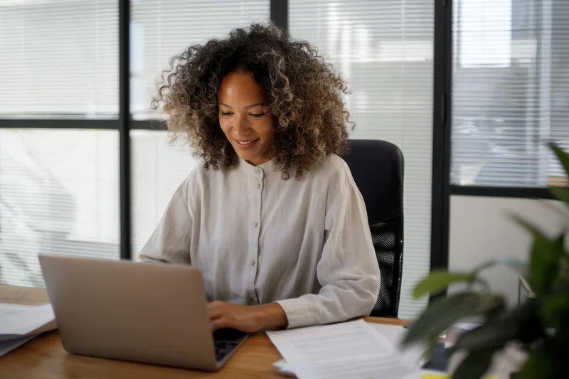 Woman working on laptop at wooden desk in office with blinds and documents nearby