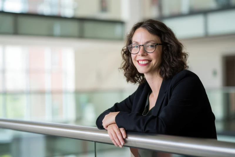 Woman in black blazer smiling while leaning on metal railing in modern office interior
