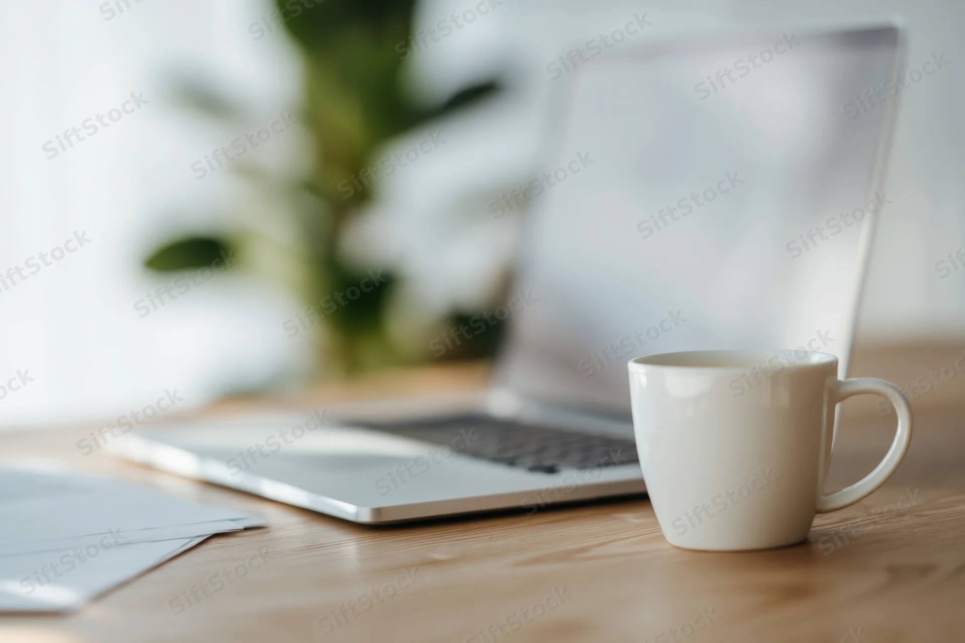 White ceramic coffee mug on wooden desk beside open laptop with blurred plant in background