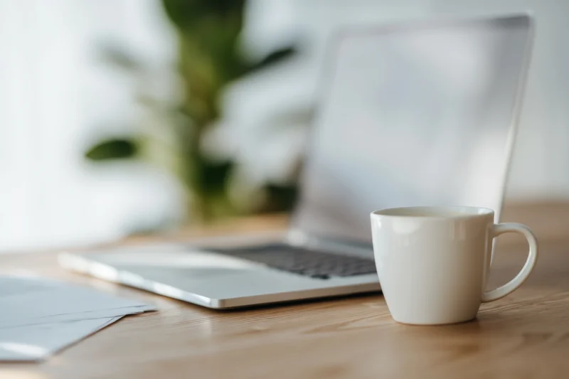 White ceramic coffee mug on wooden desk beside open laptop with blurred plant in background