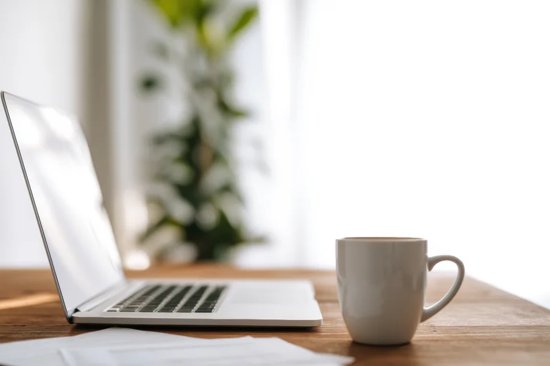 Open laptop and white ceramic mug on wooden table near window with plant in background