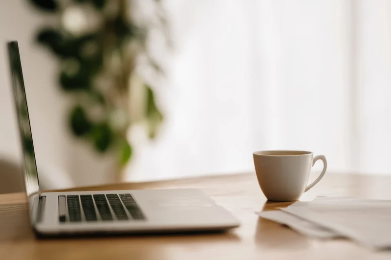 Open laptop and white ceramic coffee mug on wooden table near window with plant in background