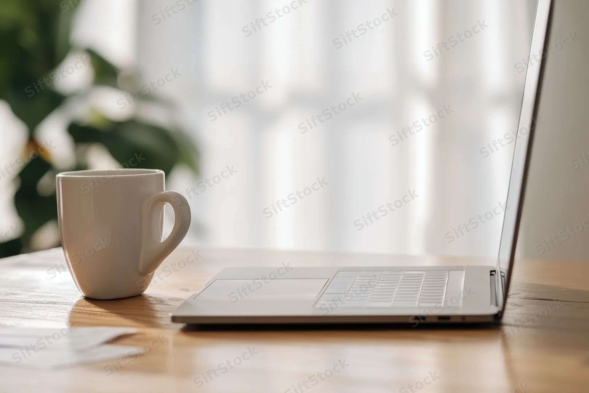 Open laptop and white ceramic coffee mug on wooden table near window