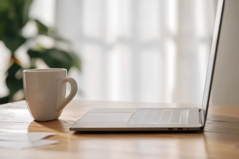 Open laptop and white ceramic coffee mug on wooden table near window