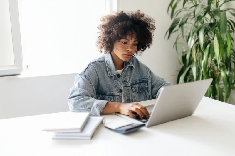 Young woman with curly hair typing on laptop at white desk in bright office space