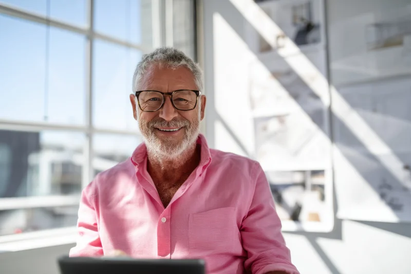 Smiling senior man with gray hair and beard wearing pink shirt in modern office workspace