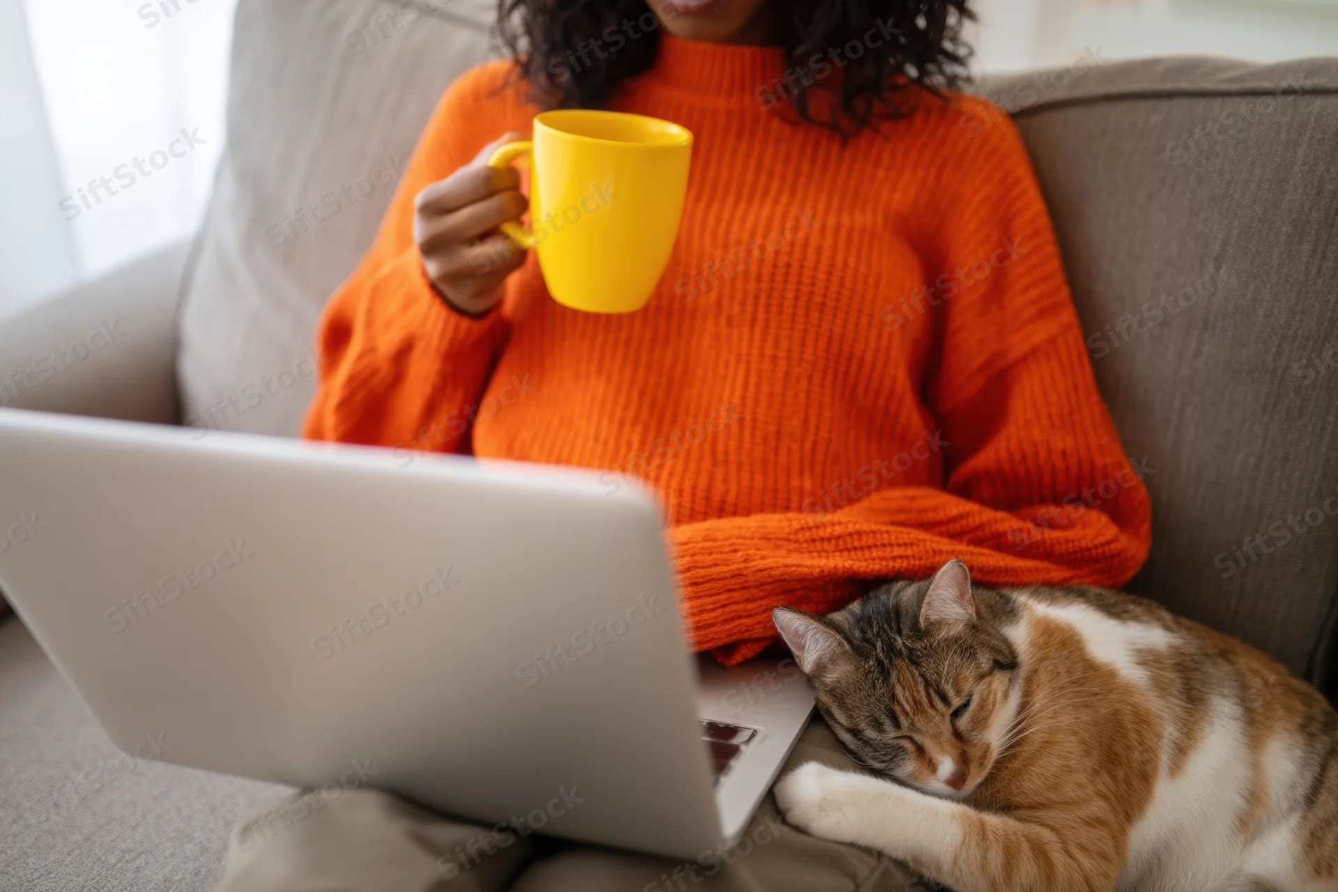 Woman in orange sweater working on laptop with cat sleeping on her lap on couch