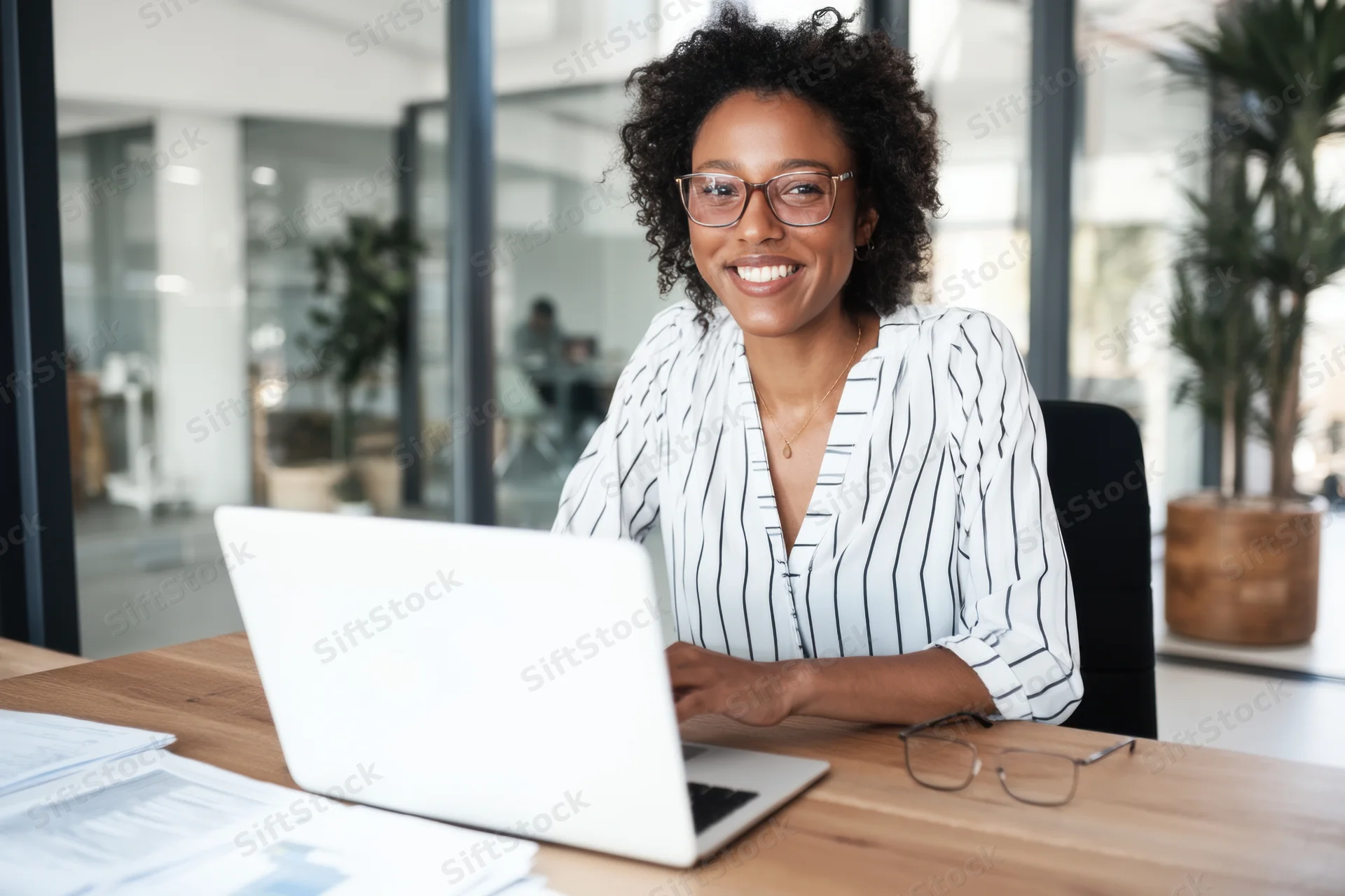 Woman smiling at camera while working on laptop at wooden desk in modern office