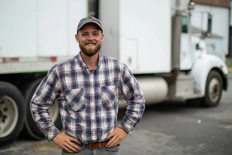 Smiling man in plaid shirt and cap standing with hands on hips in front of a white semi truck