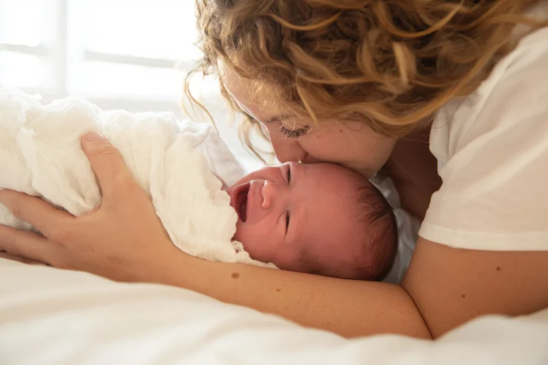 Mother kissing newborn wrapped in white blanket on bed