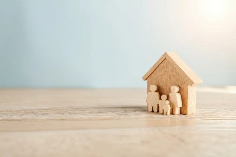 Wooden family figures standing beside a miniature house on a light wooden surface