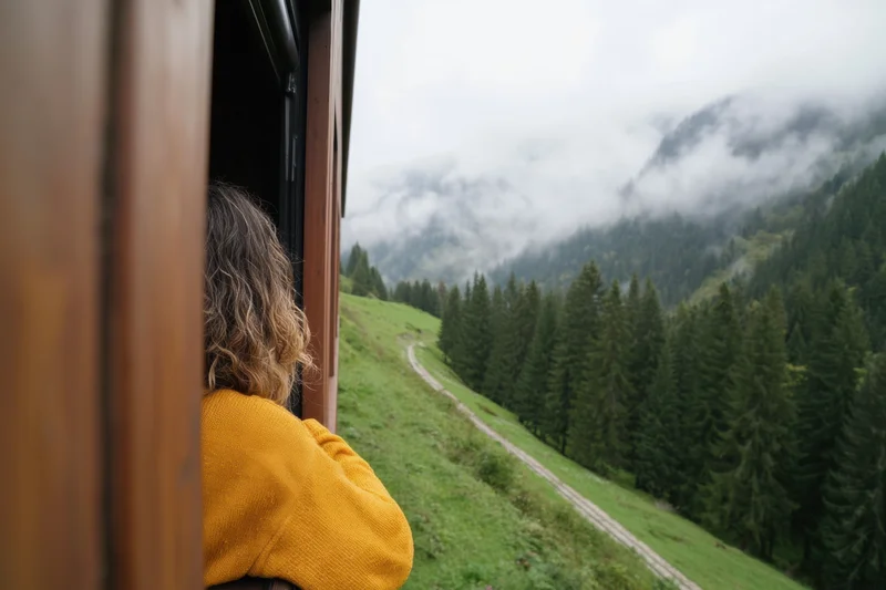 Traveler leaning out of wooden train carriage window overlooking misty mountain forest landscape