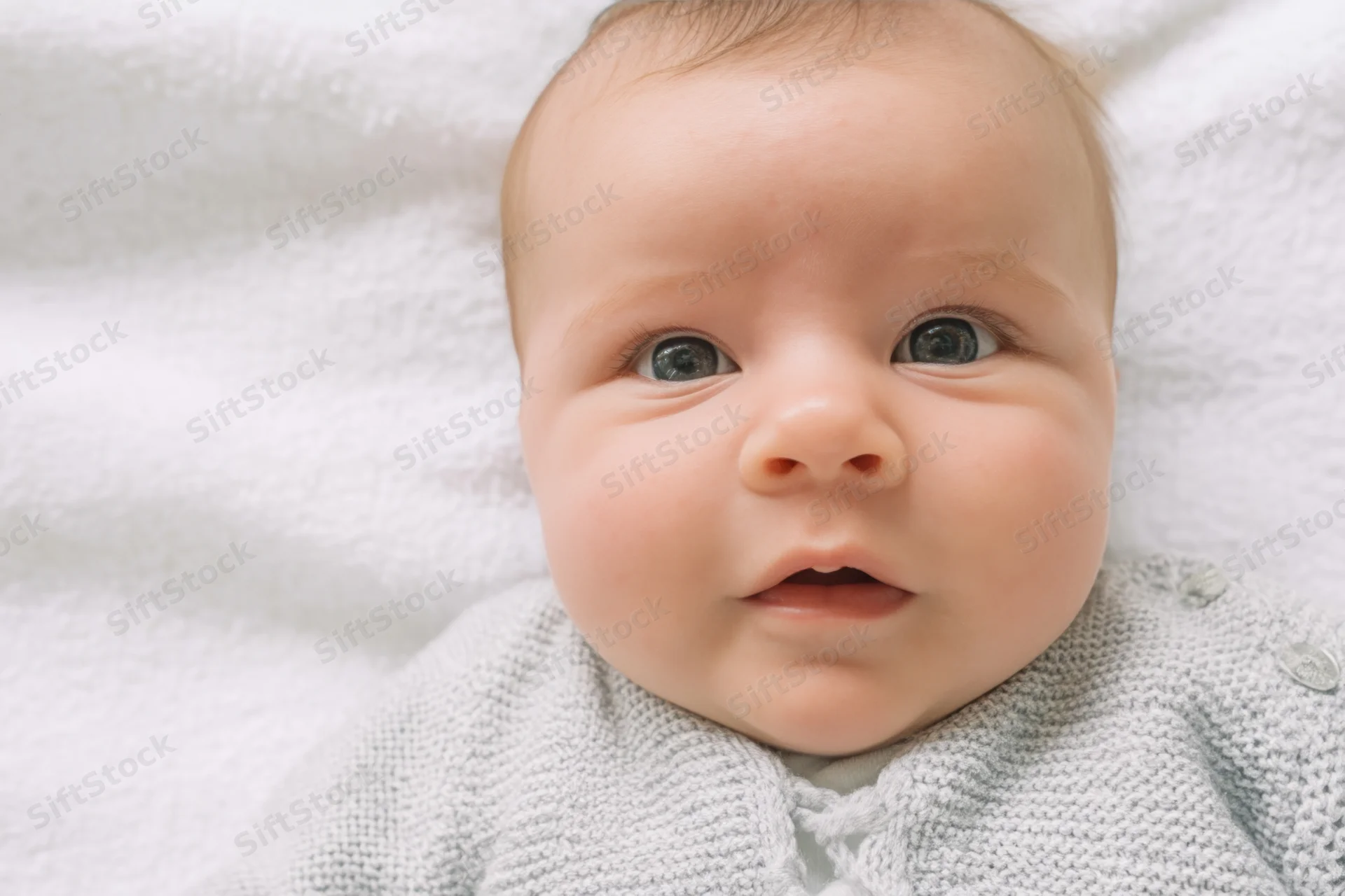 Baby lying on white blanket wearing gray knit sweater, close-up portrait of face with open mouth and blue eyes