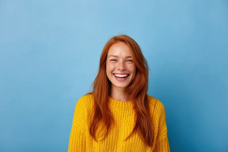 Woman smiling with long red hair wearing yellow knit sweater against blue background