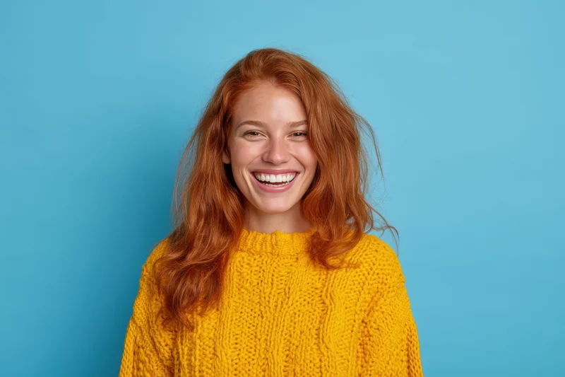 Smiling woman with long red hair wearing a yellow knit sweater against a blue background