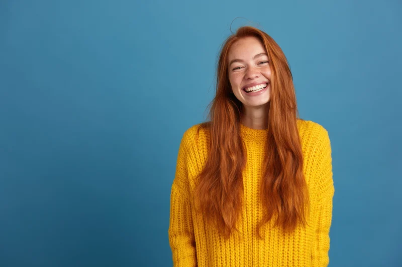 Cheerful woman with long red hair smiling in a yellow knit sweater against a blue background