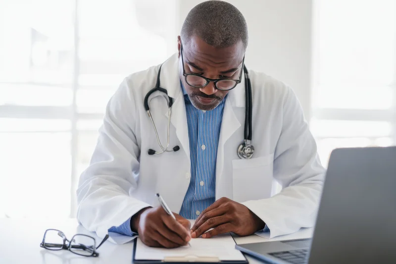 Doctor in white lab coat writing notes on clipboard at desk with laptop and stethoscope around neck