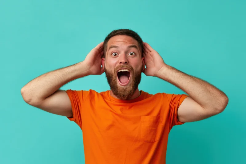 Man with beard in orange t-shirt holding head in surprise against turquoise background