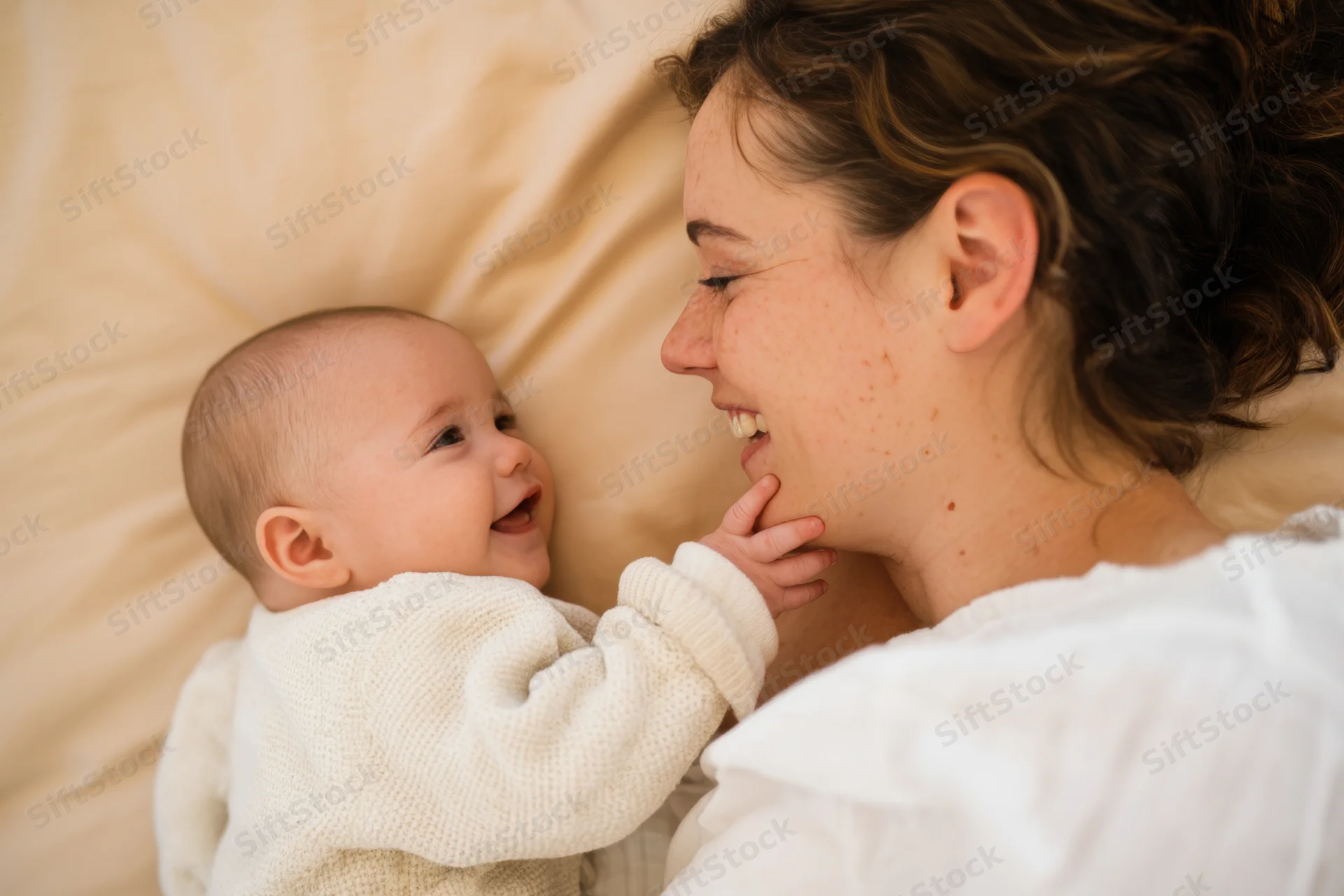 Mother smiling at her baby lying on a bed, baby touching her face