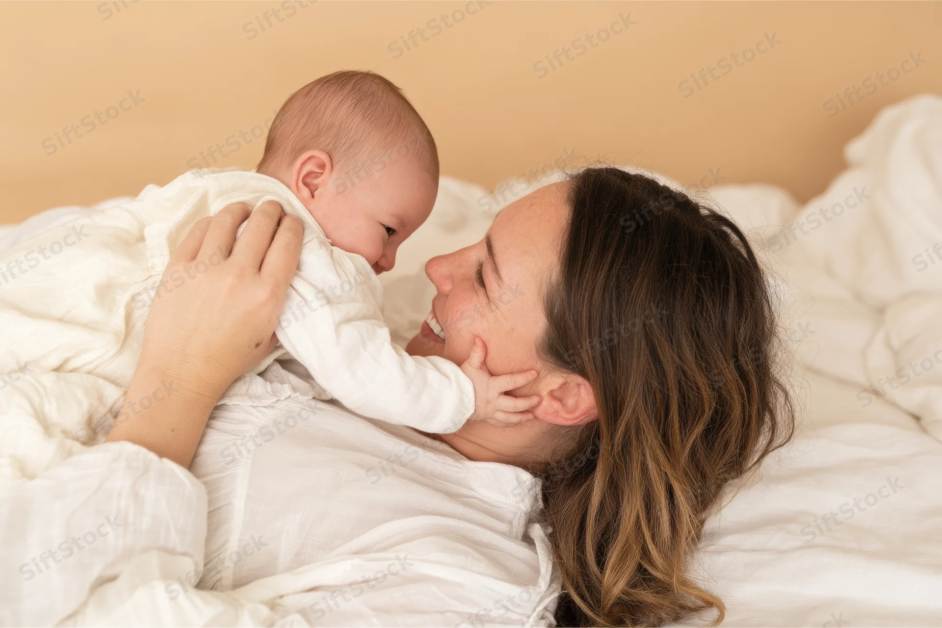 Mother smiling at her baby lying on bed in white bedding