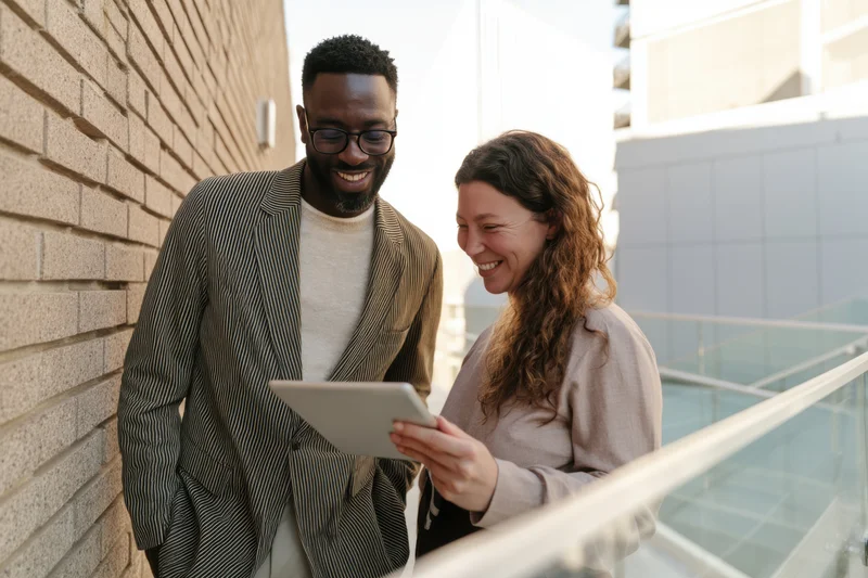 Man and woman smiling while looking at tablet together near brick wall and glass railing