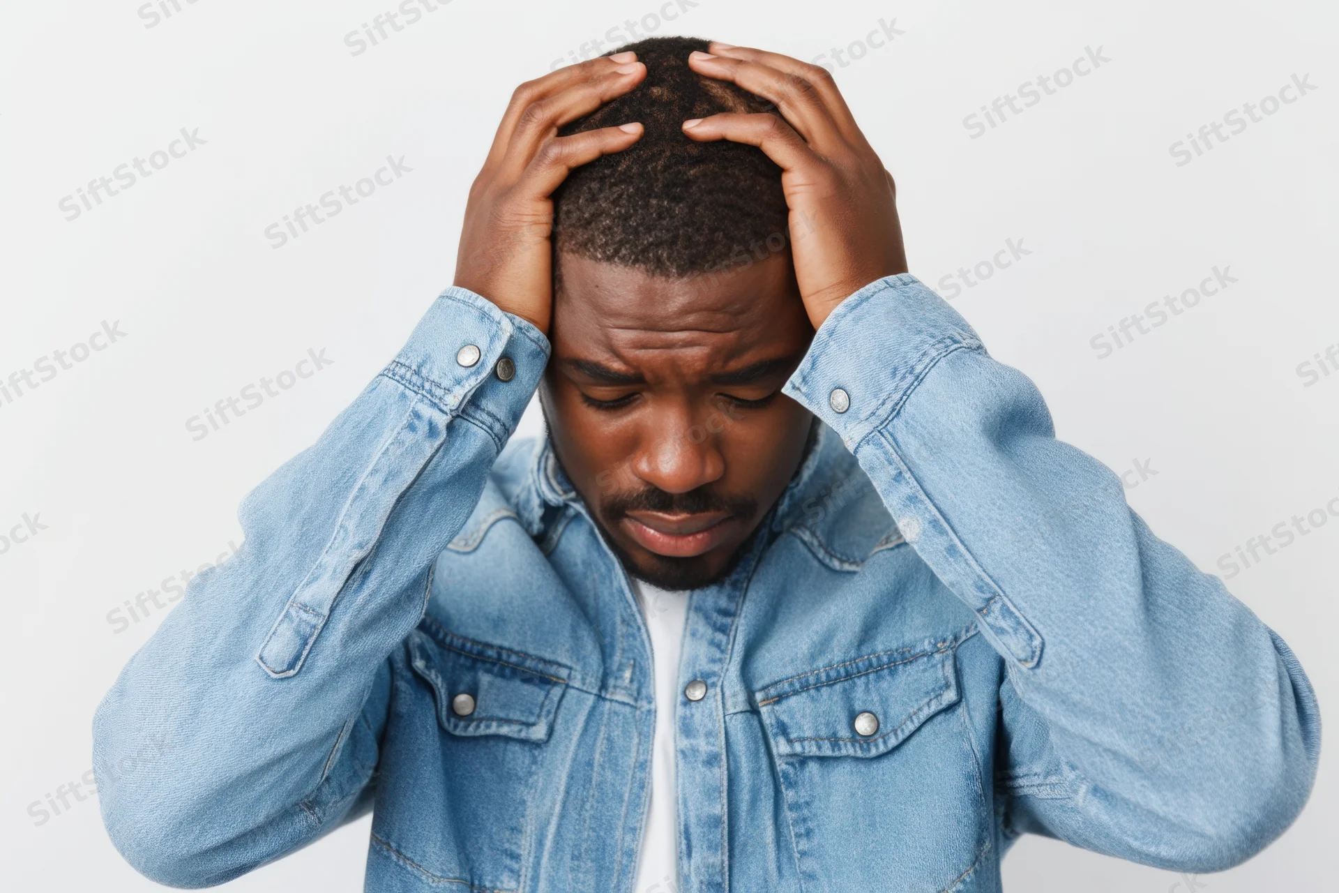 Man holding his head in distress wearing a light blue denim jacket against a white background