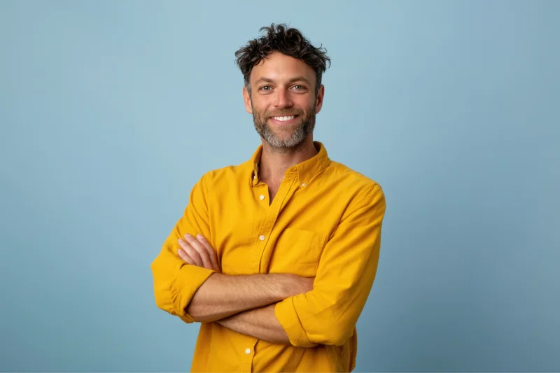Man with arms crossed smiling in yellow shirt against light blue background