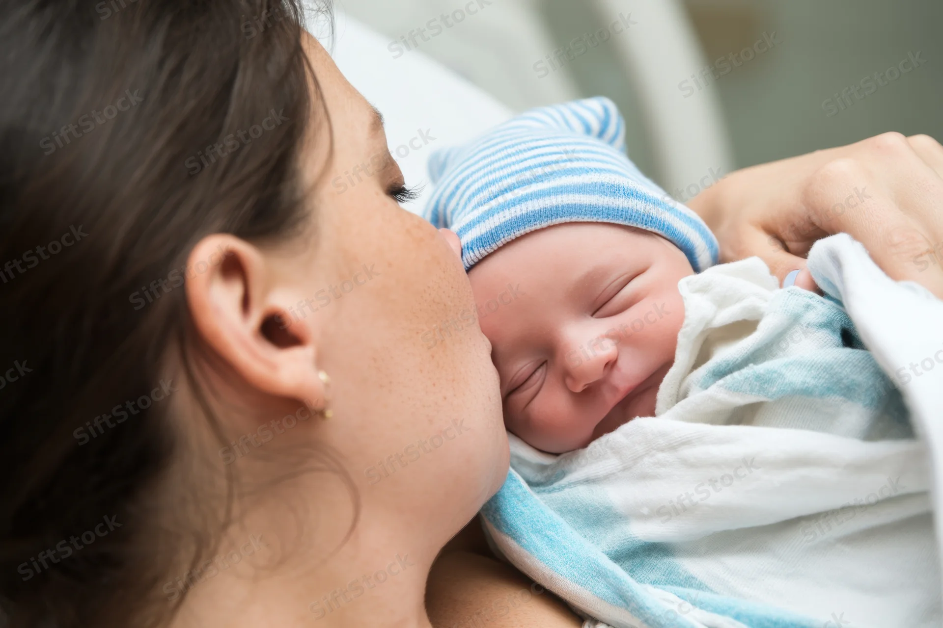 Mother kissing newborn baby wrapped in striped blanket while holding it close in hospital setting