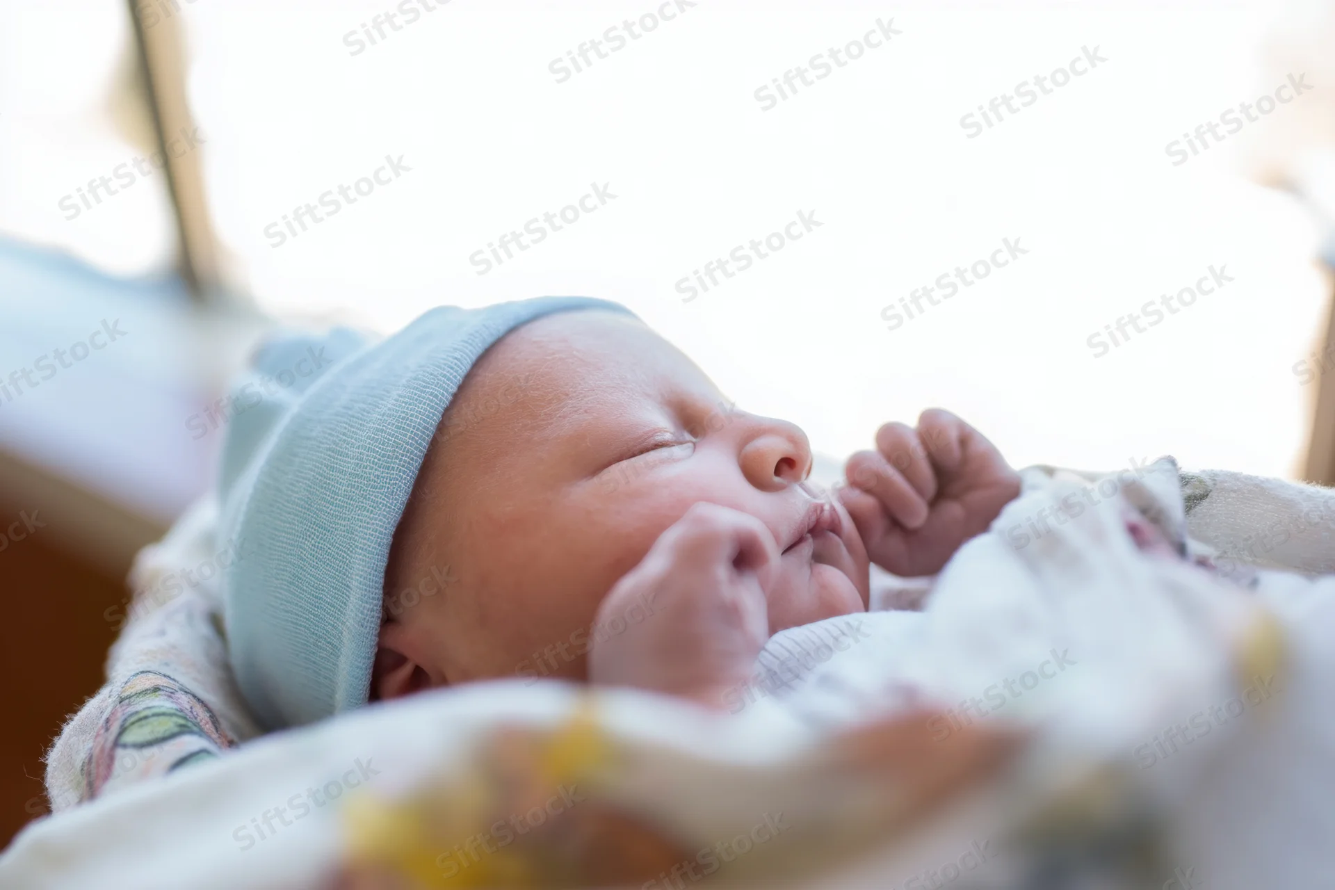 Newborn baby sleeping peacefully with eyes closed, wearing a light blue knitted hat, wrapped in a soft blanket near a window